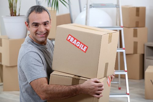 A smiling male house removals professional in casual grey clothing is indoors, holding two cardboard boxes stacked one on top of the other, with the top box displaying a red 'FRAGILE' sticker. The room appears to be a temporary storage or moving area, featuring several other packed cardboard boxes in the background, some stacked on the floor and others placed on a step ladder and shelving unit. The environment is well-lit, with neutral-toned walls and a wooden floor, indicative of a typical residential interior in the Paddington area. The presence of boxes and moving equipment, such as a step ladder, aligns with house removal services based in London. The scene emphasizes careful handling of household items during a professional move, showcasing the process of packing or unpacking in a home setting, relevant to the services provided by Paddington Man with Van on their 'Health and Safety policy' webpage.