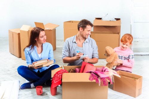 A family of three, consisting of a woman, a man, and a young girl, sit on the floor of a bright, empty room surrounded by numerous open and closed cardboard storage boxes, some filled with household items. The woman, dressed in a light blue sweater and dark blue jeans, holds a stack of books, while the man, wearing a gray shirt and dark trousers, smiles and writes on a notepad, possibly planning a move. The young girl, with red hair and a pink sweater, is holding a large teddy bear. The scene suggests they are in the process of packing or unpacking during a house move in an urban area, with the room's plain white walls and light wooden flooring providing a neutral backdrop. Several boxes in the background are stacked or partially closed, and a few items like a red mug and pink scarf are visible. This setting is typical of a residential move involving household belongings, demonstrating a typical scene of packing for house removals or transition, with a focus on both packing supplies and family involvement, aligned with professional services like Paddington Man with Van in the London area, particularly around the postcode district of Paddington.