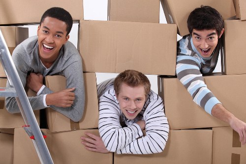 Three young men with diverse appearances are smiling and playing inside a large cardboard moving box, which is arranged among several other boxes and a metal step ladder. The man on the left, with short black hair and a grey sweater, leans on the edge of the box, showing a cheerful expression. The man in the middle, with short blond hair and a white and navy striped shirt, is lying across the boxes with his arms crossed over the edge, smiling at the camera. The man on the right, with dark, slightly curly hair and a blue and white striped shirt, is leaning into the scene from the right side, reaching out with one hand and smiling enthusiastically. The background features more stacked cardboard boxes, suggesting an ongoing packing or moving process in a residential setting, possibly in a house in Paddington or nearby London, as part of a professional removals service by Paddington Man with Van. The lighting is bright and natural, highlighting the friendly and lively atmosphere of the scene, which emphasizes the cheerful and practical nature of house relocations and packing activities.