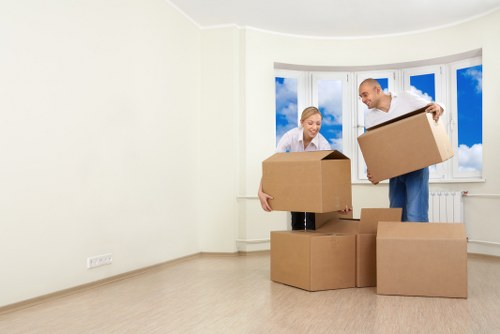 A man and a woman, both with light skin and dressed in casual clothing, are inside a bright, empty living room with beige walls and a large bay window revealing a blue sky with scattered clouds. The man is holding a cardboard box filled with various household items, and the woman is carrying a similar box. Several additional cardboard boxes are placed on the light wooden floor between them, some stacked and others positioned individually, indicating an active move. The room appears clean and well-lit, with natural daylight streaming through the window, suggesting it is daytime. The scene captures the process of packing or unpacking during a house removal, consistent with services offered by Paddington Man with Van in the local area near Paddington or Westminster, London. The background provides an unobstructed view of the outside sky, emphasizing the residential setting, and the overall scene reflects a professional and organized approach to house removals involving household furniture, boxes, and packing materials.