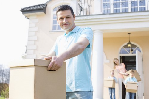A young man with dark hair, wearing a light blue polo shirt, is standing outside a cream-colored residential house with white architectural details and a porch. He is smiling and holding a cardboard moving box with both hands, appearing to be in the process of a house move. In the background, a woman and two children, all dressed casually, are also carrying cardboard boxes towards the house, indicating a family relocation. The house features a front door with an arched window above it, a hanging outdoor light, and a garden area. The scene is set in daylight with natural lighting, suggesting a typical day for house removals in the Paddington or nearby area. The image emphasizes the professional and organized nature of removals services, likely provided by Paddington Man with Van, as part of their house relocation offerings.