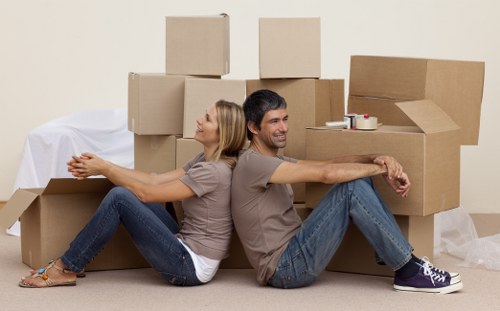 A man and a woman are sitting on the floor of a room in a relaxed pose, with their backs against various cardboard moving boxes stacked behind them. The boxes are of different sizes, some closed and others partially open, showing packing material and household items inside. The woman, with blonde hair, wears a light beige t-shirt, blue jeans, and sandals, while the man, with dark hair, wears a grey t-shirt, blue jeans, and sneakers. The room has a plain light-colored wall and carpeted flooring. In the background, a white sheet covers some larger objects or furniture, and there are rolls of tape and packing supplies placed on one of the boxes. The scene suggests a domestic setting in the process of a house move, typical of a house removal service in the Paddington area, with clear visual focus on the packed belongings and the casual, content atmosphere of the movers taking a break amid the packing effort.