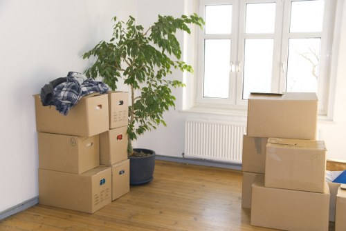 Image depicts a bright, tidy room with white walls and a wooden floor, featuring several cardboard moving boxes stacked on the left and right sides of the space. On the left, a small stack of three boxes with printed symbols and labels is topped with a folded piece of fabric, possibly a blanket or cloth. To the right, a larger box is stacked atop another, with additional boxes partially visible at the bottom of the frame. A tall potted plant with broad green leaves is positioned near the left corner, adding a natural element to the scene. In the background, a large window with four panes allows natural light to fill the room, and a white radiator is mounted directly underneath it. The overall setting suggests an interior preparing for a house removal or transition, with minimal furniture or personal items visible, consistent with a professional removals service environment in the Paddington area. The space appears clean and organized, ready for packing or loading, reflecting the typical stage of a house move managed by Paddington Man with Van in London.