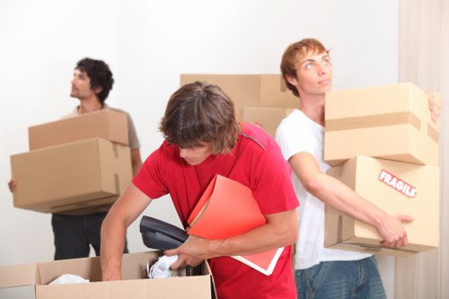 Three men involved in house removals are inside a residential room with white walls. The central figure, wearing a red shirt, is leaning over a large cardboard box, holding a black telephone handset in his right hand and carrying a red folder along with some papers in his left arm. Behind him, a man with dark curly hair, dressed in a grey t-shirt, is carrying a medium-sized cardboard box in each hand, with a serious expression and looking away. To the right, a young man with light brown hair, wearing a white t-shirt, is holding two medium-sized cardboard boxes, one in each hand, with a label indicating 'Fragile'. The boxes are of plain brown cardboard, with some having visible tape seals. The scene appears to be set in a home environment, possibly a hallway or living room, with minimal background details. The lighting is natural and even, emphasizing the focus on the men and their packing activities during the house removal process, as part of a professional moving service in Paddington or nearby areas in London.
