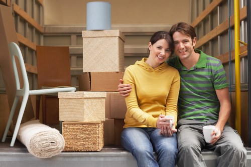 A smiling young couple sits closely together on a step inside a storage unit, surrounded by various packed moving boxes and household items. The woman, wearing a yellow hoodie and blue jeans, holds a white mug in her hands, while the man, dressed in a green and purple striped polo shirt and grey trousers, also holds a mug. Behind them, there are stacked square cardboard boxes, some covered with protective blankets or woven baskets, with a tall beige storage box topped by a light blue container in the background. To the left, a light grey chair is partially visible, and a rolled-up white rug lies on the floor nearby. The environment appears to be a wooden-floored storage space or garage, with wooden shelving and beams framing the scene. Natural, soft lighting illuminates the area, evoking a sense of preparation and transition associated with house removals in London or nearby areas, subtly reflecting the services of Paddington Man with Van, a local removals company. The overall scene suggests a cheerful moment during a moving process, with household items organized for transportation or storage.