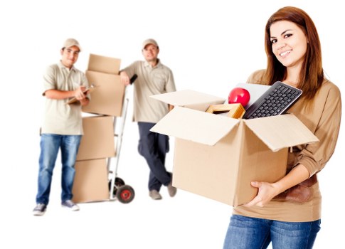 A young woman with long brown hair, dressed casually in a tan top and blue jeans, is smiling and holding a large open cardboard box filled with various household items, including a computer keyboard, a red apple, and a bottle. In the background, two male movers, dressed in light-colored work shirts, are standing on a bright, neutral backdrop. One is holding a box and a mobile device, while the other is standing next to a red and black hand truck loaded with stacked moving boxes. The scene suggests a house removal or relocation process, with the furniture and boxes indicating an indoor residential setting, possibly within a property in the Paddington or London area. The lighting is even and natural, emphasizing the professional yet friendly atmosphere of the house removals service offered by Paddington Man with Van, known for assisting home relocations in the local vicinity.