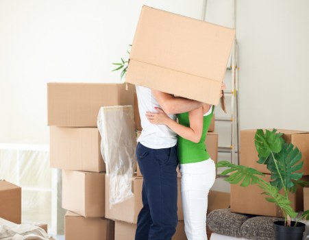 In a well-lit residential interior, two individuals are engaged in a house removals process, with one person lifting a medium-sized cardboard box, their arms and hands visible, partially obscured by the box. The other person, standing behind, is supporting the box with one arm around it, their torso visible, dressed in a white top and dark trousers. Surrounding them are several stacked cardboard boxes of varying sizes, some sealed and others open with packing material visible. The background shows a plain white wall, part of a wooden staircase, and a beige carpeted floor. To the right, a black pot containing a large, lush green plant with broad leaves adds a natural touch, while a small upholstered stool is partially visible. The scene depicts a typical moment during a residential move within London or nearby areas, supporting the house removals services offered by Paddington Man with Van, with natural daylight illuminating the space, emphasizing the organized packing and preparation for a move.