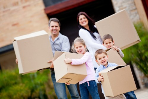 A family of five, including two adults and three children, standing outdoors in front of a residential property with a brick and stone exterior, engaging in a house move. The family members are holding large cardboard boxes, which are plain brown with visible flaps and tape. The adults are smiling, one woman with dark hair wearing a white top and a man in a light-colored shirt, both positioned behind the children. The children, a girl and two boys, are happily holding boxes, with the girl in a pink top and the boys in casual clothing. The woman appears to be carrying or balancing a box, while the other adult is also holding a box. The background features a driveway, some greenery, and part of the house's exterior wall, which includes brickwork and stone detailing. The scene is well-lit with natural daylight, capturing a moment typical of household removals or moving services in the London area, subtly referencing Paddington or nearby locations. The overall image conveys a positive, organized move involving family members and basic packing supplies, relevant to house removals services offered by Paddington Man with Van.