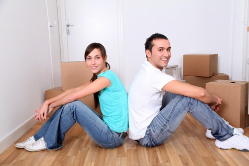 A young man and woman sitting back-to-back on a light wooden floor inside a residential property, surrounded by cardboard moving boxes in various sizes, some stacked and others placed on the floor. The woman, wearing a turquoise t-shirt and blue jeans, has long brown hair and is smiling at the camera. The man, dressed in a white t-shirt and blue jeans, also smiles and looks slightly to the right. The background features a plain white wall with a closed white door and minimal decor, indicating the interior of a house in the Paddington area, suitable for local removals and house moving services by Paddington Man with Van. The natural lighting highlights the clean, uncluttered space, emphasizing the preparation phase of a house relocation, with items ready to be packed or unloaded during a typical move on a residential street in London postcode area W2, close to Paddington station.