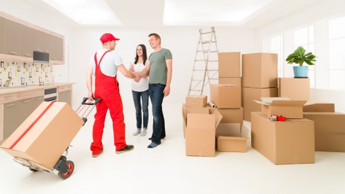 In a well-lit, modern residential interior, a house removal scene is depicted with a removals technician dressed in red overalls and a red cap, engaged in a handshake with a smiling couple. The technician is holding a small hand trolley with a few packed moving cartons, while in the background, a large metal ladder leans against the wall near a window, suggesting preparations for a move within the property. Multiple cardboard boxes of varying sizes, some sealed with packing tape and others open revealing contents, are stacked neatly across the floor, indicating packing activities typical of house removals in the Paddington or central London area. A potted plant with broad green leaves sits on a windowsill, providing a touch of home comfort. The scene captures the organized, professional environment of house moving services, emphasizing the safety and care taken during a domestic relocation process, with bright natural daylight illuminating the space and highlighting the clean, uncluttered interior perfect for household moves in northwest London.