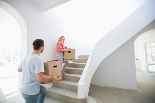 A young woman with blonde hair, dressed in a pink checkered shirt and light-colored pants, is carrying a cardboard box up a flight of white, curved staircase in a bright, modern house interior. A man with short dark hair, wearing a light blue t-shirt, is standing at the bottom of the stairs, holding another cardboard box. The staircase has a smooth, white banister and is situated in an open, well-lit hallway with large windows allowing natural daylight to fill the space. The flooring appears to be light grey or speckled, complementing the clean and contemporary aesthetic of the residential property, suggesting a professional house removal or relocation scenario in the Paddington area or nearby London postcode. The background shows a spacious, minimalist interior with white walls and a glass-paneled door leading to additional rooms, emphasizing the home environment where Paddington Man with Van offers removal services, including packing and transportation of household belongings.