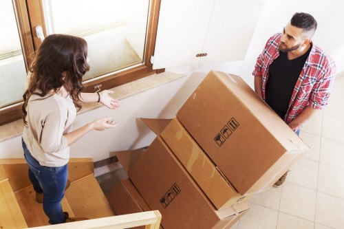A man and a woman are engaged in a conversation inside a residential property, with the man holding three large, sealed cardboard boxes, likely packed for a house move in the Paddington area. The woman, standing on a wooden staircase, gestures towards the boxes as they discuss their contents or the moving process. The scene is set near a window with a white radiator beneath, allowing natural light to illuminate the space, which features a light-colored tiled floor and white walls. The cardboard boxes are standard brown with printed handling symbols on their sides, indicating careful handling during relocations. The man is dressed casually in a plaid shirt and dark trousers, while the woman wears a light sweater and blue jeans. The setting appears to be a hallway or entrance area of a typical London flat or house, with the interior designed for practical use during a house removals service by Paddington Man with Van, reflecting the standard procedures for loading household belongings prior to transportation.