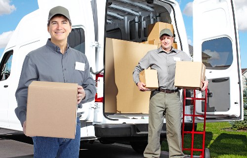 Two professional removal workers in grey uniforms and grey caps are shown in front of a white moving van parked on a residential street in Paddington. The worker on the left is smiling and holding a large cardboard box, while the worker on the right is carrying a similar box and standing beside a red dolly. The open rear doors of the van reveal stacked cardboard boxes, some of which are sealed and others partially visible, along with the interior space prepared for loading household belongings. The background features a well-maintained green lawn and a hint of housing buildings. The scene depicts a professional house removal service, with clear details of cardboard boxes, the van, and the workers preparing for a house move, emphasizing the logistics and support provided by Paddington Man with Van services in the local area.