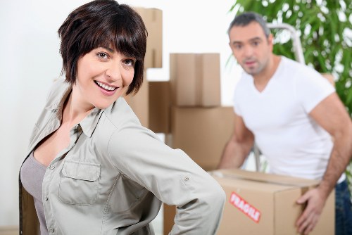 A woman with short dark hair smiling at the camera while standing in a well-lit room filled with cardboard moving boxes, some stacked in the background. She is wearing a light grey work jacket, indicating involvement in a house removals or packing process. Behind her, a man with short dark hair and a beard, dressed in a white t-shirt, is slightly out of focus and appears to be lifting or moving a cardboard box, suggesting active loading or unloading. The setting is inside a residential space, possibly an entrance hall or living room, with natural light illuminating the scene and a green plant visible to the right. Several packed boxes of various sizes are arranged around the scene, some with labels or tape, emphasizing a storage or relocation context. The overall environment reflects a professional removals company preparing to assist with household moves near Paddington, consistent with local house removal services focused on sustainability and recycling.