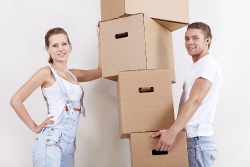 A young woman and a young man are standing indoors in a residential setting, each holding cardboard boxes during a house removal process. The woman, positioned on the left, is wearing light blue denim overalls over a white tank top, with a friendly expression and her right arm resting on an open box while her left hand supports her hip. The man, on the right, is dressed in a white T-shirt and light blue jeans, smiling as he holds a closed cardboard box at waist height with both hands. Behind them, several stacked cardboard boxes are arranged vertically, some open and others closed, suggesting packing or unpacking activities typical of a house move. The background features a plain, light wall, and the lighting appears natural, highlighting the texture of the cardboard boxes and their casual clothing, fitting naturally within the context of local house removals and relocation services in Paddington. The scene emphasizes organized packing, familiar household items, and a professional yet relaxed atmosphere associated with professional removals companies like Paddington Man with Van.