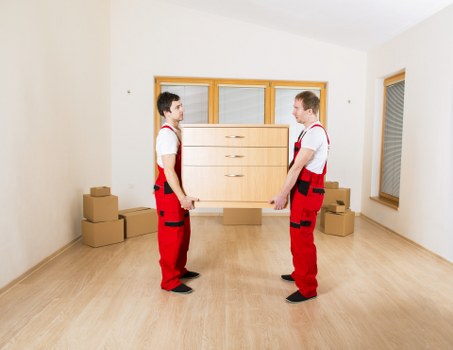 Two movers in red work trousers and white shirts are standing in a bright, empty room with light wooden flooring and white walls, each holding a side of a large wooden dresser with multiple drawers. The dresser has a natural wood finish and is being held horizontally between them. In the background, there is a large window with blinds on the right side, and behind the movers, there are several stacked cardboard boxes placed on the floor, likely for house removals or relocation purposes. The room appears to be a minimally furnished interior, possibly in a residential property within the Paddington area, with ample natural light coming through the window, creating a clear and professional scene. The setting emphasizes the careful handling and transportation of household furniture typical of Paddington Man with Van’s house removals services in London.