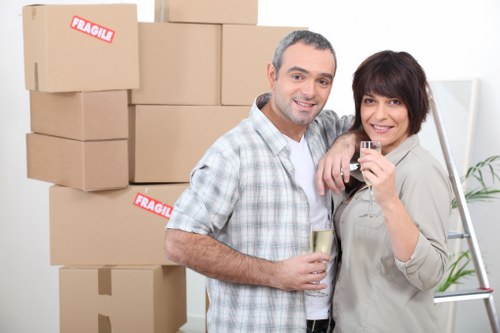 A man and woman stand inside a residential property, smiling at the camera while holding glasses of champagne or sparkling wine, suggesting a celebration or farewell during a house move. Behind them, there are several large cardboard moving boxes with red 'FRAGILE' stickers, stacked against a plain white wall. The boxes vary in size and appear to be made of brown corrugated cardboard. The man has short, grey hair and is dressed in a short-sleeved, checked shirt, while the woman has short, dark hair styled in a layered cut and is wearing a light-colored jacket. To the right, a metallic step ladder is partially visible, indicating recent packing or unpacking activity. The setting appears to be a clean, well-lit living room or hallway, with a glimpse of a potted plant in the background, enhancing the domestic atmosphere. This image, associated with Paddington Man with Van’s house removals services in London, subtly highlights the preparation and celebration involved in relocating homes within the Paddington area, with a focus on professionalism and careful handling of household belongings.