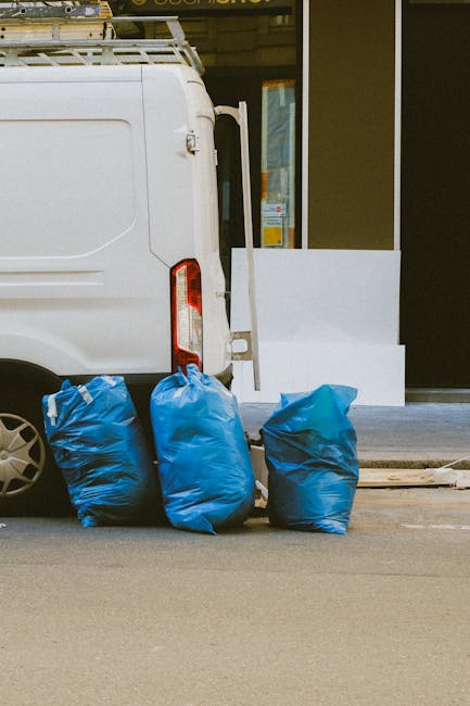 An outdoor scene showing a large, weathered green waste disposal container filled with various discarded items including cardboard boxes, plastic packaging, and paper waste. The container is situated 