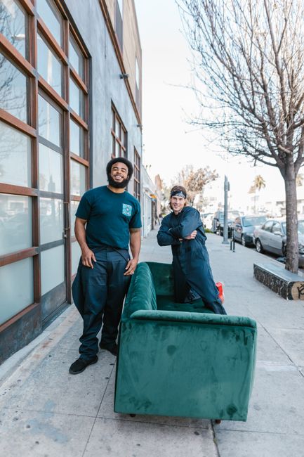 Two movers from [COMPANY_NAME] stand on a residential sidewalk in W2, London, during a home relocation process. One mover, an African descent man with a beard, wears a navy blue T-shirt and dark trous