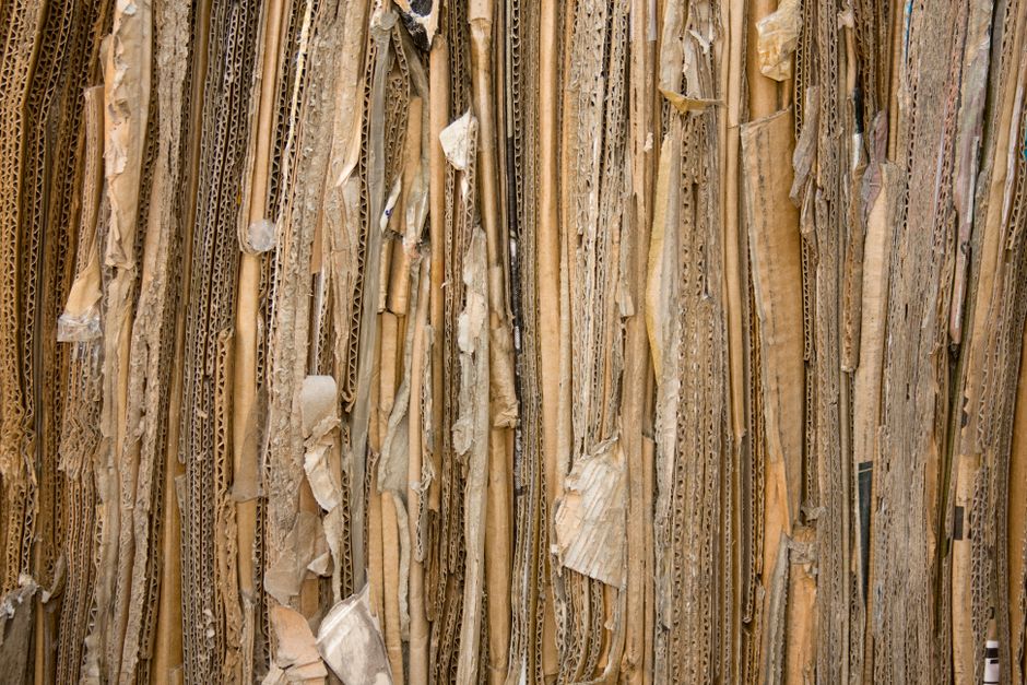 Close-up view of a stack of flattened cardboard boxes and packing materials, including corrugated cardboard and paper sheets, leaning against a wall in an interior space. The materials are bundled tog