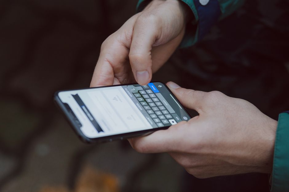 Close-up of a person's hands holding a smartphone, which displays a calculator and some text related to moving costs or quotes. The person is outdoors, with a blurred natural background, and is wearin
