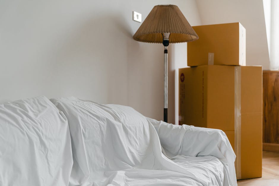 Indoor scene showing a person lying on the floor amidst a collection of cardboard moving boxes of various sizes, some sealed with packing tape and others open or partially assembled, indicating an act